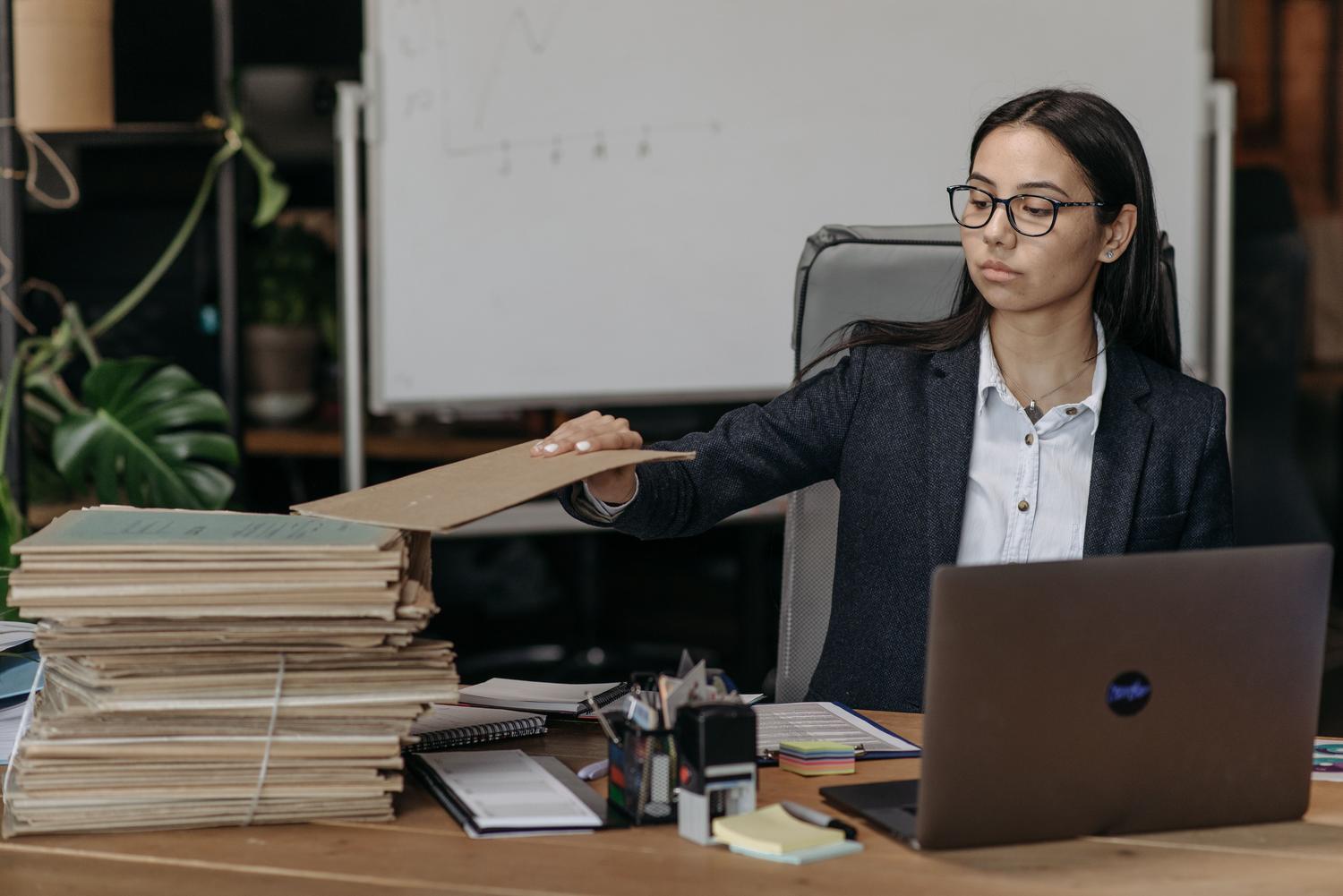 Mujer de negocios gestionando una gran pila de carpetas físicas en una oficina, ilustrando el caos de organizar miles de archivos manualmente.