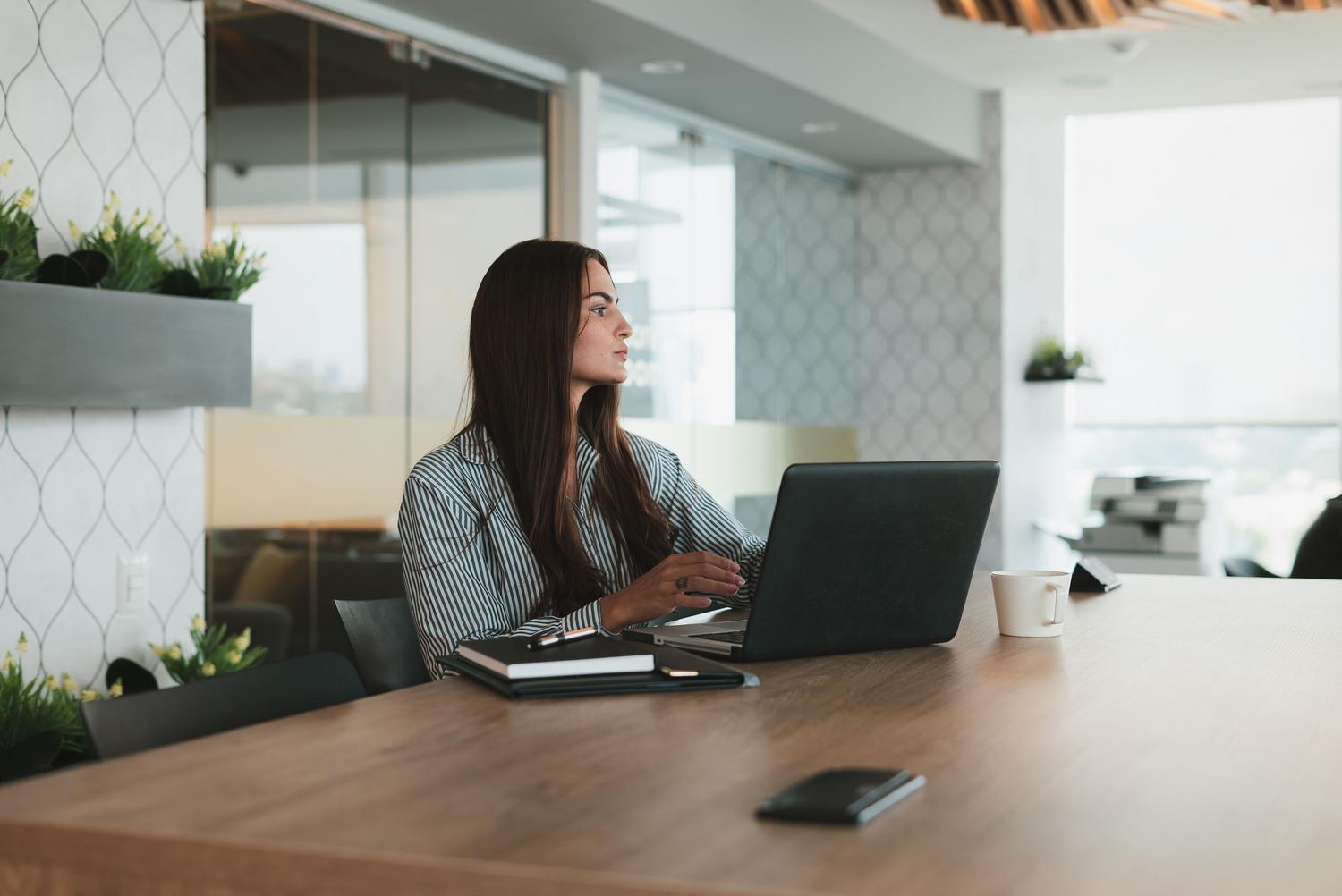 Mujer de negocios reflexionando frente a su ordenador en una oficina luminosa y moderna, ilustrando el proceso de toma de decisiones y brainstorming con herramientas de IA.