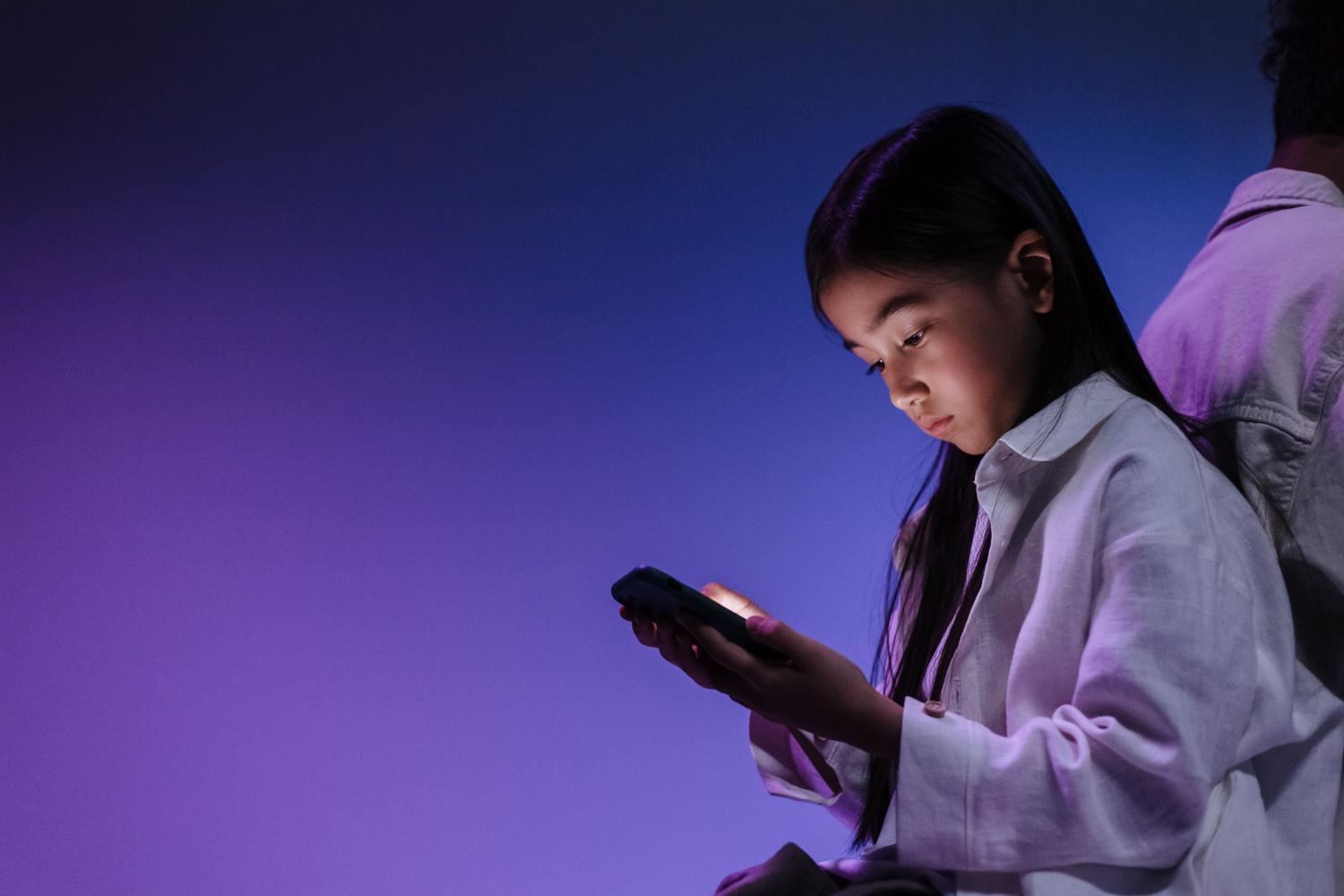Niña absorta en su teléfono móvil bajo una luz ambiental azul y púrpura, representando la inmersión digital y los riesgos de las redes sociales.